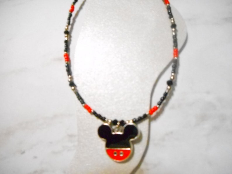 Pode incluir: Um colar de contas com um pingente do Mickey Mouse. O colar tem contas pretas e vermelhas com detalhes dourados. O pingente do Mickey Mouse &eacute; preto com uma base vermelha e uma borda prateada.