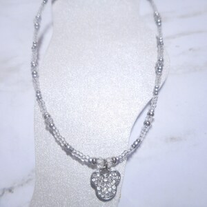 May include: A silver anklet featuring a Mickey Mouse charm encrusted with clear rhinestones. The anklet is strung with clear beads and small silver beads. The charm is approximately 0.75 inches wide.