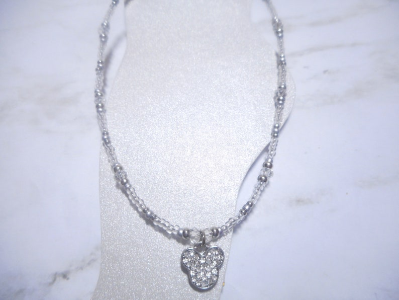 May include: A silver anklet featuring a Mickey Mouse charm encrusted with clear rhinestones. The anklet is strung with clear beads and small silver beads. The charm is approximately 0.75 inches wide.