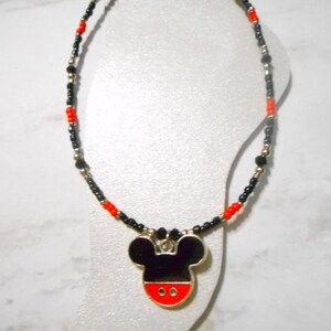 Pode incluir: Colar de contas com um pingente do Mickey Mouse. O colar tem contas pretas, vermelhas e prateadas. O pingente &eacute; preto com uma base vermelha e contorno prateado. Apresentado em um manequim branco.
