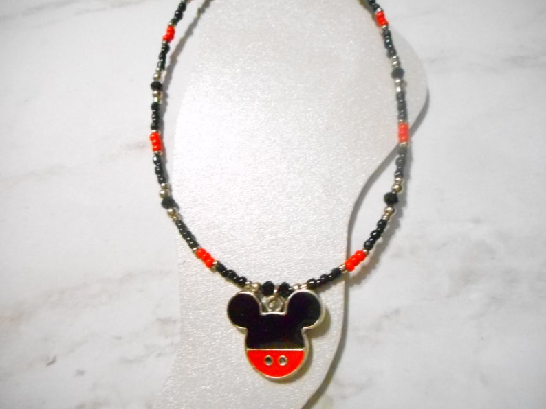 Pode incluir: Colar de contas com um pingente do Mickey Mouse. O colar tem contas pretas, vermelhas e prateadas. O pingente &eacute; preto com uma base vermelha e contorno prateado. Apresentado em um manequim branco.