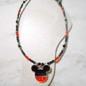 Pode incluir: Um colar de contas com um pingente do Mickey Mouse. O colar tem contas pretas, vermelhas e prateadas. O pingente &eacute; preto com base vermelha e acabamento prateado. O colar &eacute; exibido em um manequim branco.