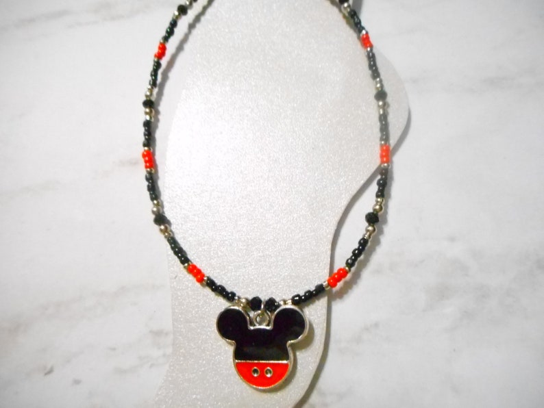 Pode incluir: Um colar de contas com um pingente do Mickey Mouse. O colar tem contas pretas, vermelhas e prateadas. O pingente &eacute; preto com base vermelha e acabamento prateado. O colar &eacute; exibido em um manequim branco.