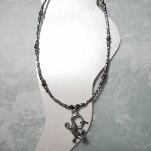 May include: A beaded anklet with a silver-colored lizard pendant. The anklet features small, dark beads and larger, faceted beads. The lizard pendant is detailed and adds a unique touch to the jewelry. The anklet is displayed on a white foot-shaped form.