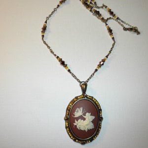 Op de afbeelding: Een bruine cameo-hangerketting met een wit vlinder- en bloemenmotief. De ketting is gemaakt van metaal en heeft een bruin-gouden ketting met kleine kralen.