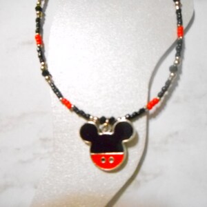 Pode incluir: Um colar com um pingente do Mickey Mouse. O pingente &eacute; preto, vermelho e branco com uma borda prateada. A corrente do colar &eacute; feita de contas pretas, vermelhas e douradas.
