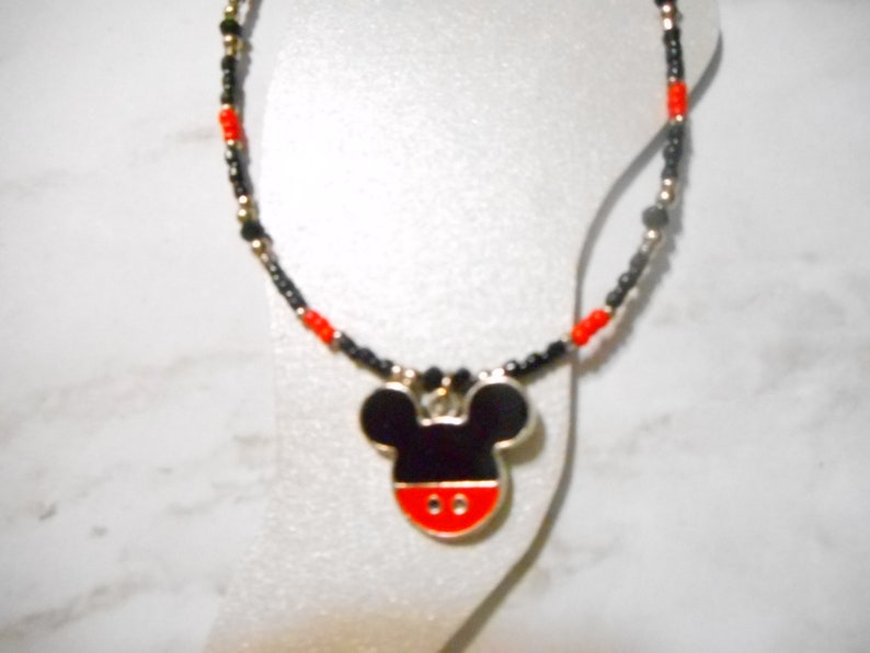 Pode incluir: Um colar com um pingente do Mickey Mouse. O pingente &eacute; preto, vermelho e branco com uma borda prateada. A corrente do colar &eacute; feita de contas pretas, vermelhas e douradas.