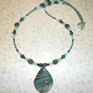 May include: A green agate pendant necklace with silver wire wrapped around the stone. The necklace is made with green and white beads and a silver clasp.