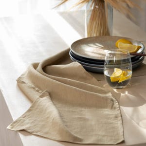 May include: A beige linen table runner draped across a light-colored surface. A stack of plates, a glass of water with lemon slices, and a vase with dried plants are in the background. The scene is lit with natural light, creating a soft, neutral aesthetic.
