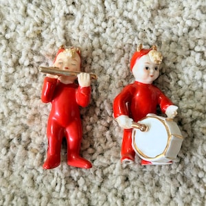May include: Two porcelain figurines of figures dressed in bright red suits with gold-trimmed hoods. One figure plays a gold flute with eyes closed, while the other figure plays a white drum with gold trim, featuring a gold drumstick in hand.