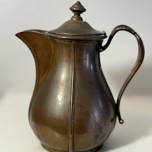 May include: A vintage bronze-colored pitcher with a curved spout and a handle. The pitcher has a lid with a decorative finial. The body of the pitcher has vertical lines. The pitcher is likely made of metal and is a decorative item.