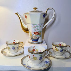 May include: A vintage tea set featuring a cream-colored teapot and three teacups with saucers. The set is adorned with floral patterns in red, blue, and yellow, with gold trim. The teapot is approximately 10 inches tall.