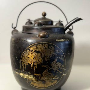 Puede incluir: Tetera antigua negra y dorada con asa y boquilla curvadas. La tetera presenta una ilustración dorada de una grulla y árboles dentro de un diseño circular. La tapa tiene un pequeño pomo y dos pomos más pequeños.