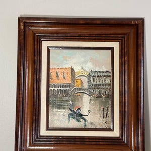 Könnte beinhalten: Ölgemälde einer venezianischen Kanalszene, gerahmt in dunklem Holz. Das Kunstwerk zeigt eine Gondel, Brücken und Gebäude mit orangefarbenen Dächern. Der Himmel ist eine Mischung aus Blau und Weiß, und das Wasser spiegelt die Architektur wider.
