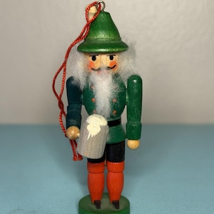 May include: A wooden Christmas ornament shaped like a nutcracker. It wears a green jacket, black shorts, and orange trousers. The figure has a white beard, a green hat, and holds a beer mug. A red string is attached for hanging, suitable for a Christmas tree.