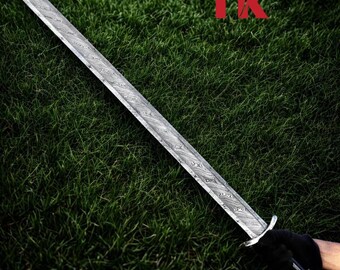 Op de afbeelding: Een lang, zilveren zwaard met een patroon op het lemmet, tegen een achtergrond van groen gras. De handgreep en de pareerstang zijn ook zilver. Het lemmet heeft een uniek, getextureerd ontwerp. De letters "TK" zijn rood.