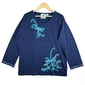 May include: A long-sleeved navy blue pullover shirt featuring embroidered turquoise and light blue dragonfly and vine motifs. The neckline and sleeve cuffs are accented with decorative stitching. A white brand label is visible at the inner neck.