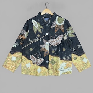 May include: A dark denim jacket featuring a button-down front, a collar, and long sleeves. The garment is decorated with embroidered butterfly and leaf motifs in shades of green, brown, and white, with a yellow patterned panel along the hem.