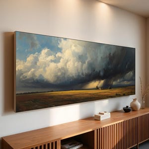 May include: A large rectangular landscape painting depicting a dramatic sky with dark storm clouds and bright sunlight over a field. The artwork is framed and mounted on a white wall above a wooden cabinet.