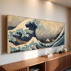 May include: A framed print of Hokusai's "The Great Wave off Kanagawa" features a large, curling wave in shades of blue and white, with Mount Fuji in the background. The artwork is displayed on a wall above a wooden cabinet.