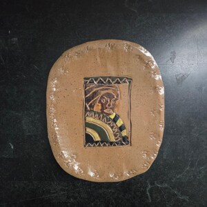 May include: A handmade, oval-shaped ceramic plate with a central rectangular image of a person. The image features a person with a yellow and black striped garment. The plate is tan with a textured surface.
