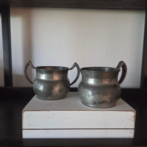 Peut inclure: Deux crémier en étain antiques avec des poignées incurvées. Les crémier ont une forme bombée et reposent sur un bloc rectangulaire blanc. L'arrière-plan est une étagère sombre et un mur blanc. Les crémier ont un aspect vieilli et terni.