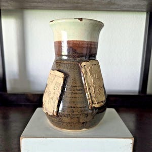 Könnte beinhalten: Eine handgefertigte Keramikvase mit einzigartigem Design. Die Vase hat einen hellgrünen Rand, ein braunes Band und einen dunkelbraunen Körper mit zwei strukturierten, goldfarbenen rechteckigen Akzenten. Sie steht auf einem weißen quadratischen Sockel.