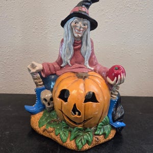 Könnte beinhalten: Eine Halloween-Dekorationsfigur aus Keramik. Sie zeigt eine lächelnde Hexe mit schwarzem Hut, die auf einem geschnitzten orangefarbenen Kürbis sitzt. Die Hexe hält einen roten Apfel und wird von einem Totenkopf und blauen Stiefeln flankiert.