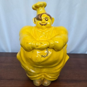 May include: A vintage ceramic cookie jar in the shape of a chef. The jar is primarily yellow with brown hair and a smiling face. The chef is wearing a tall hat and apron, with arms crossed. A chain detail hangs from the apron.