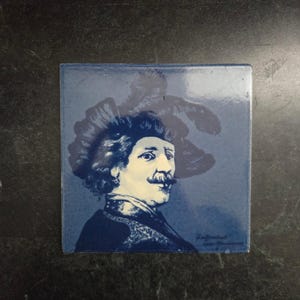 May include: Square blue tile featuring a portrait of a man with a large mustache and a feathered hat. The portrait is in a dark blue and white color scheme. The tile has a glossy finish and is likely a decorative item.