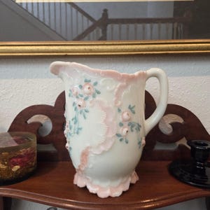 May include: A white ceramic pitcher with a pink ruffled rim and handle. The pitcher is decorated with pink roses and green leaves. The pitcher is sitting on a wooden shelf.