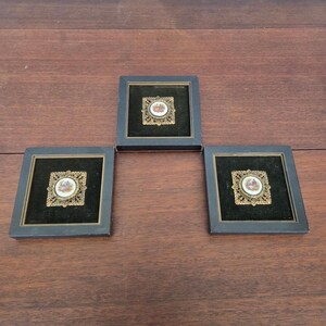 May include: Three framed miniature artworks. Each piece features a small, circular, ornate design in gold and white, set against a black velvet background. The frames are square and black, creating a classic presentation. The artworks are displayed on a dark wood surface.