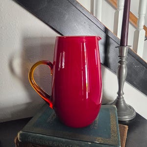 Könnte beinhalten: Eine leuchtend rote Glaskaraffe mit gelbem Griff, die auf einem Stapel antiker Bücher steht. Die glatte Oberfläche der Karaffe reflektiert das Licht, und der Griff ist elegant geschwungen. Eine dunkle Treppe und ein Kerzenhalter mit einer lila Kerze befinden sich im Hintergrund.