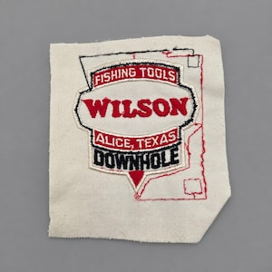 May include: A rectangular off-white fabric patch featuring an embroidered emblem with red and black text. The emblem reads FISHING TOOLS, WILSON, ALICE, TEXAS, and DOWNHOLE. A thin red embroidered outline of a geometric shape surrounds the emblem.