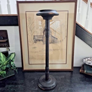May include: A tall, dark brown hat stand with a round base and a circular top. The stand is made of wood and has a decorative, turned design on the central pole. The stand is placed in front of a framed drawing.