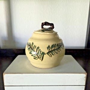 May include: A small, round, cream-colored ceramic jar with a lid and a dark brown handle. The jar is decorated with green leaf patterns. It sits on a white, rectangular box.