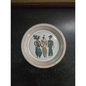 May include: A round, off-white ceramic plate with a raised rim. The plate features a hand-painted design of three figures in colorful attire. The figures are depicted in shades of green, blue, and tan. The plate has a diameter of approximately 8 inches.