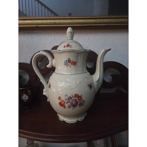 May include: Cream-colored porcelain coffee pot with a lid, handle, and spout. The pot is decorated with floral patterns in red, purple, and blue. It has a green trim and sits on a dark wooden surface.