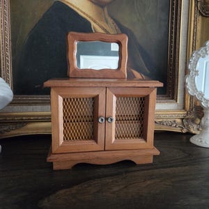 May include: A small, wooden jewelry cabinet with a rectangular mirror on top. The cabinet has two doors with wire mesh inserts and decorative knobs. The wood has a warm, natural finish. The cabinet is sitting on a dark surface.