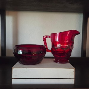 Könnte beinhalten: Ein rubinrotes Sahne- und Zuckerdosen-Set aus Glas. Der Sahnekännchen hat einen Griff und einen Ausguss, und die Schüssel hat eine abgerundete Form. Beide Teile haben ein geometrisches Muster. Das Set wird auf einer weißen Holzoberfläche präsentiert.