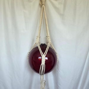 Puede incluir: Un macetero colgante de macramé con un globo de cristal rojo oscuro. El macramé blanquecino tiene un diseño anudado, que suspende el globo. El globo es una esfera, que refleja la luz. El macetero está suspendido de un anillo metálico.