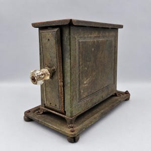 May include: A vintage metal toaster with a weathered, dark green finish and a rectangular body. It features a clear, faceted glass knob on the side, a flat top, and a sturdy base with four small feet. The metal surface shows signs of oxidation.