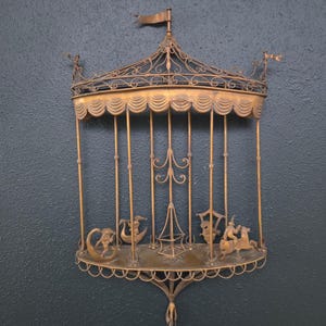 May include: Ornate, antique-style brass wall shelf. The shelf features a decorative cage design with a curved top, vertical bars, and a lower shelf with whimsical figures. A small flag sits atop the structure. The shelf is mounted on a dark blue wall.