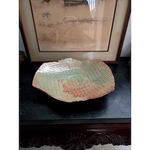 May include: A shallow, wide ceramic bowl with an irregular, wavy rim. The interior surface features a textured grid pattern in reddish-brown tones, overlaid with a smooth, glossy glaze in a pale mint green color that pools in the center.