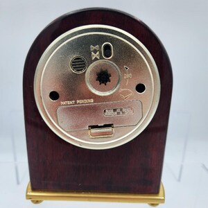 以下が含まれることがあります： 濃い茶色の木製仕上げと金色のアクセントが施された小さな時計。時計の背面には、さまざまな設定とバッテリーコンパートメントを備えた円形の金属プレートがあります。「Patent Pending」の文字が見えます。