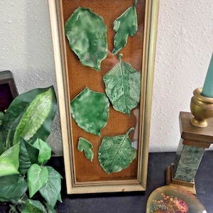 Puede incluir: Una obra de arte enmarcada con varias hojas de cerámica verde sobre un fondo de arpillera marrón. Las hojas varían en tamaño y forma, con vetas detalladas. El marco es de color oro claro, añadiendo un toque vintage a esta pieza de temática natural.