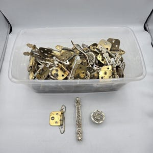 May include: A clear plastic bin filled with various brass and silver-toned cabinet hardware. Several pieces are displayed below, including a hinge, a handle with an ornate design, and a round knob. The hardware appears to be vintage or antique.