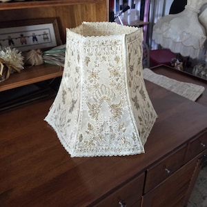May include: Cream-colored lampshade with a scalloped edge and a gold floral pattern. The lampshade has a hexagonal shape and is made of a textured fabric. It sits on a dark wooden surface.