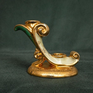 May include: A gold-colored, ornate candle holder with a cream-colored, curved body and green accents. The base and decorative swirls are gold. The candle holder has two circular openings for candles.