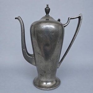 May include: A tall, metallic gray teapot with a smooth, rounded body, a slender curved spout, and a high-arched handle. The vessel features a tapered base and a removable lid topped with a small, pointed finial. The surface has a matte finish.